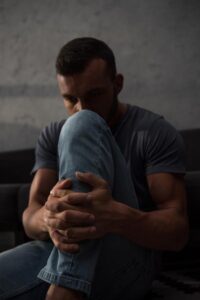 distressed looking man sitting on the floor hugging his knee with face partially obscured by knee