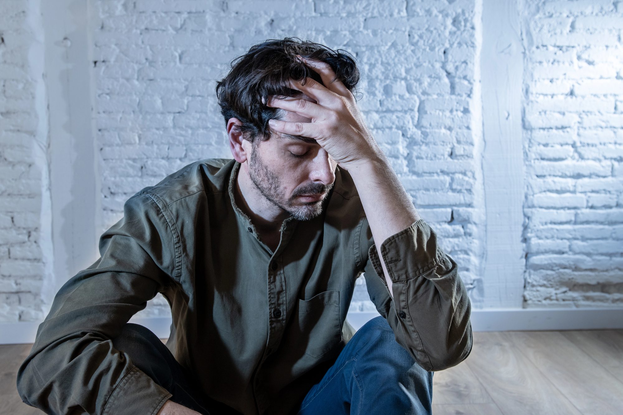 Young depressed man sitting against a white wall at home with a shadow on the wall feeling miserable, lonely and sad in mental health depression concept