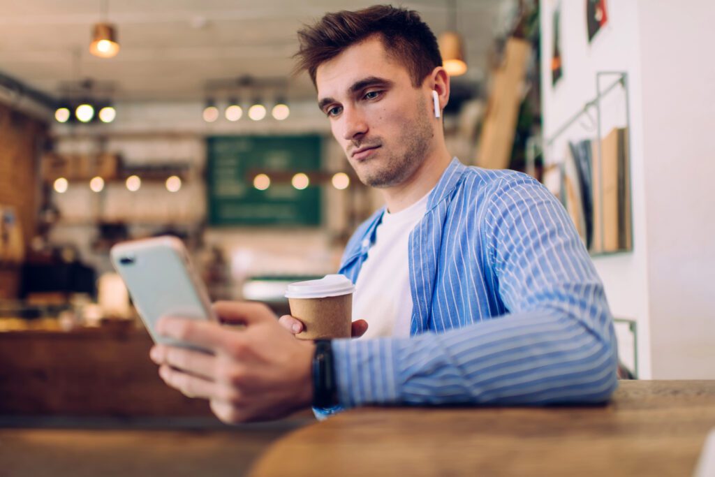 Back view of serious man with dark hair in casual clothes holding coffee and browsing smartphone while messaging with friends in cafeteria