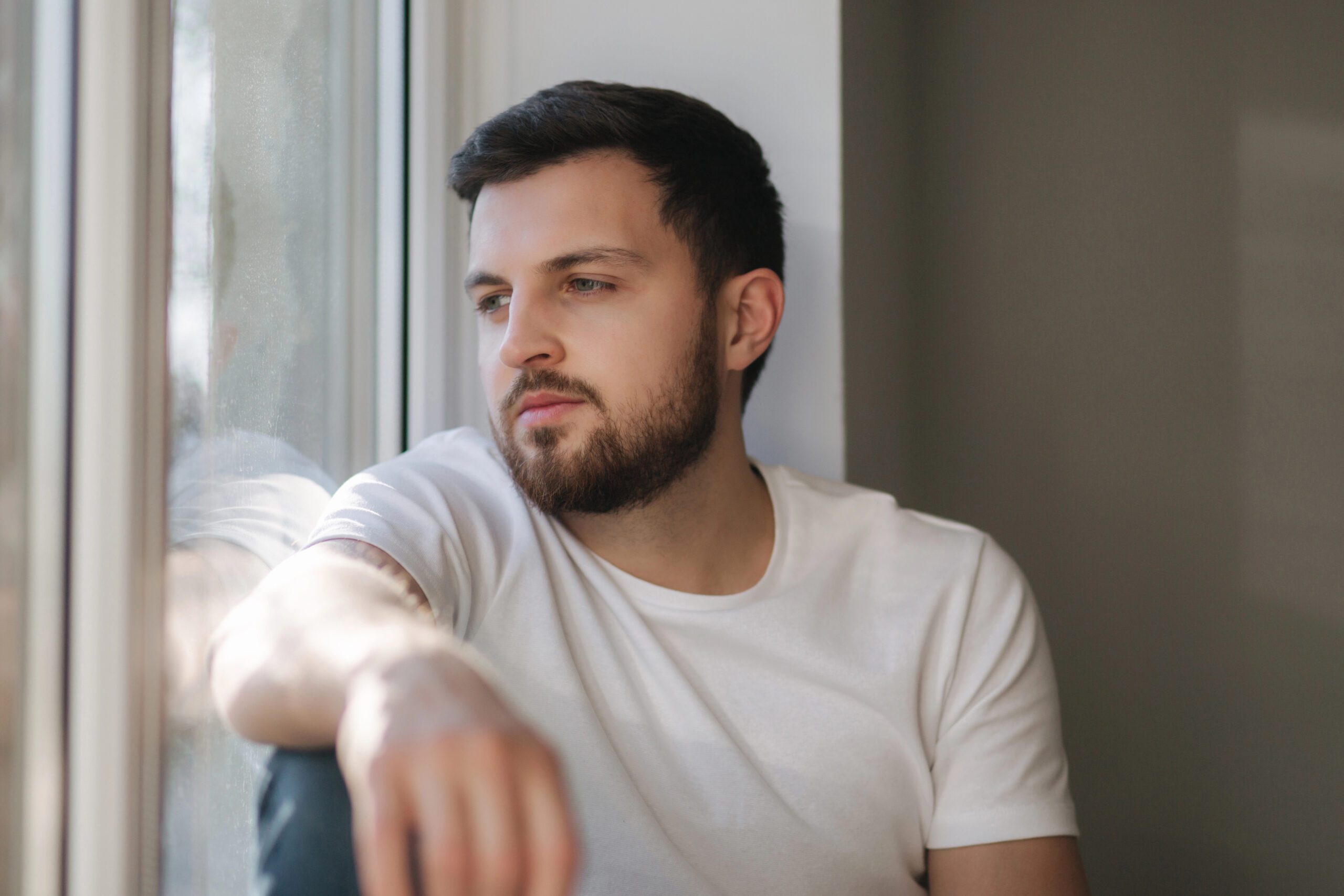 Young man sits on windowsill and looks out the window. Alone at home