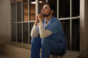 Male nurse sitting on windowsill at hospital in evening, experiencing medical professional stress