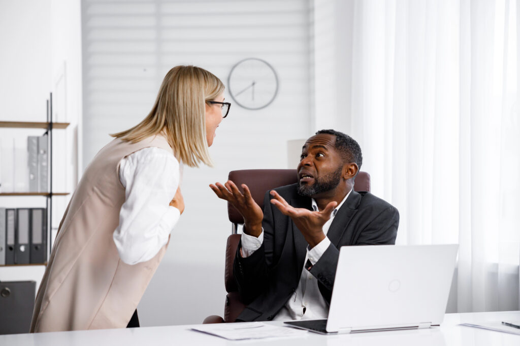 A woman leans toward a seated man and yells as he raises his hands beside an office laptop.