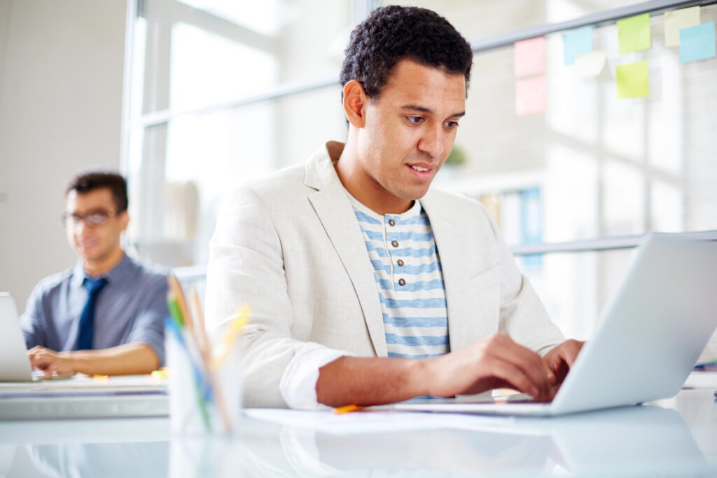 A man in a light blazer types on a laptop at his office desk.