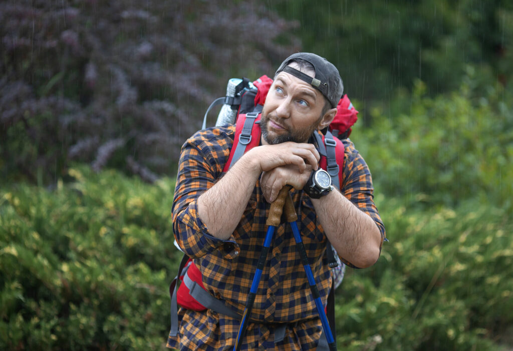 A man with a backpack rests on his trekking poles in a green forest.